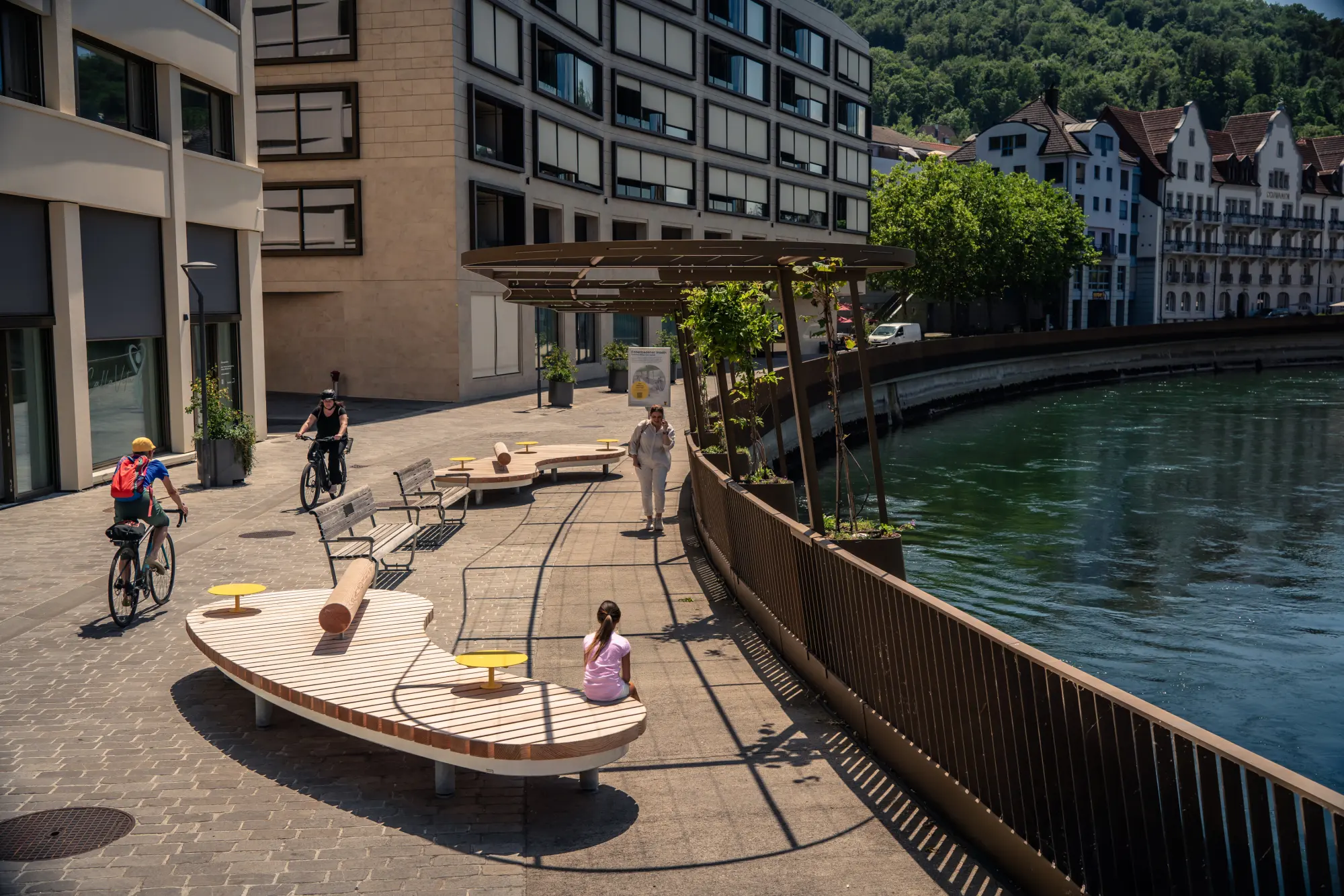 Geschwungene Holzsitzlandschaft mit Pergola entlang der Limmatpromenade Ennetbaden, Passanten und Radfahrende am Flussufer.