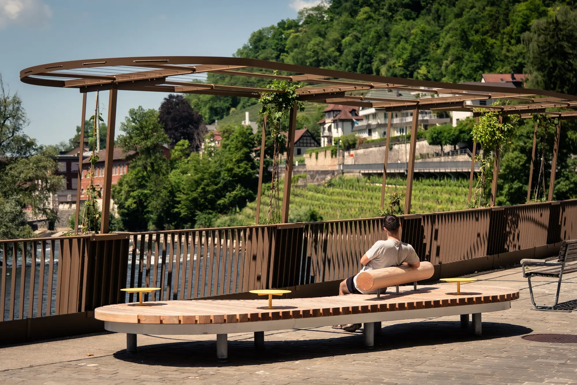 Gestaltete Sitzlandschaft mit Holzliegebank und Pergola an der Limmatpromenade Ennetbaden, Blick über den Fluss auf die Badstrasse und Weinberge.