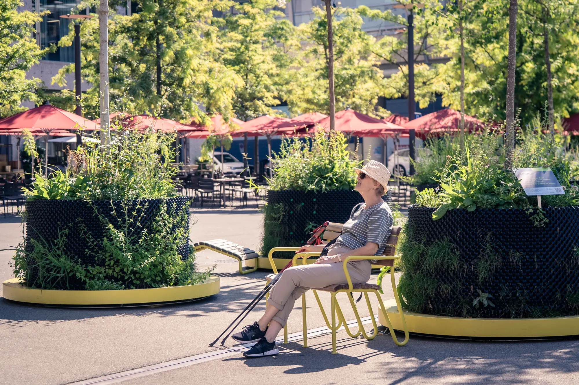 Frau sitzt entspannt zwischen mobilen Baumpflanzgefässen mit dichter Bepflanzung, im Hintergrund rote Sonnenschirme eines Straßencafés.
