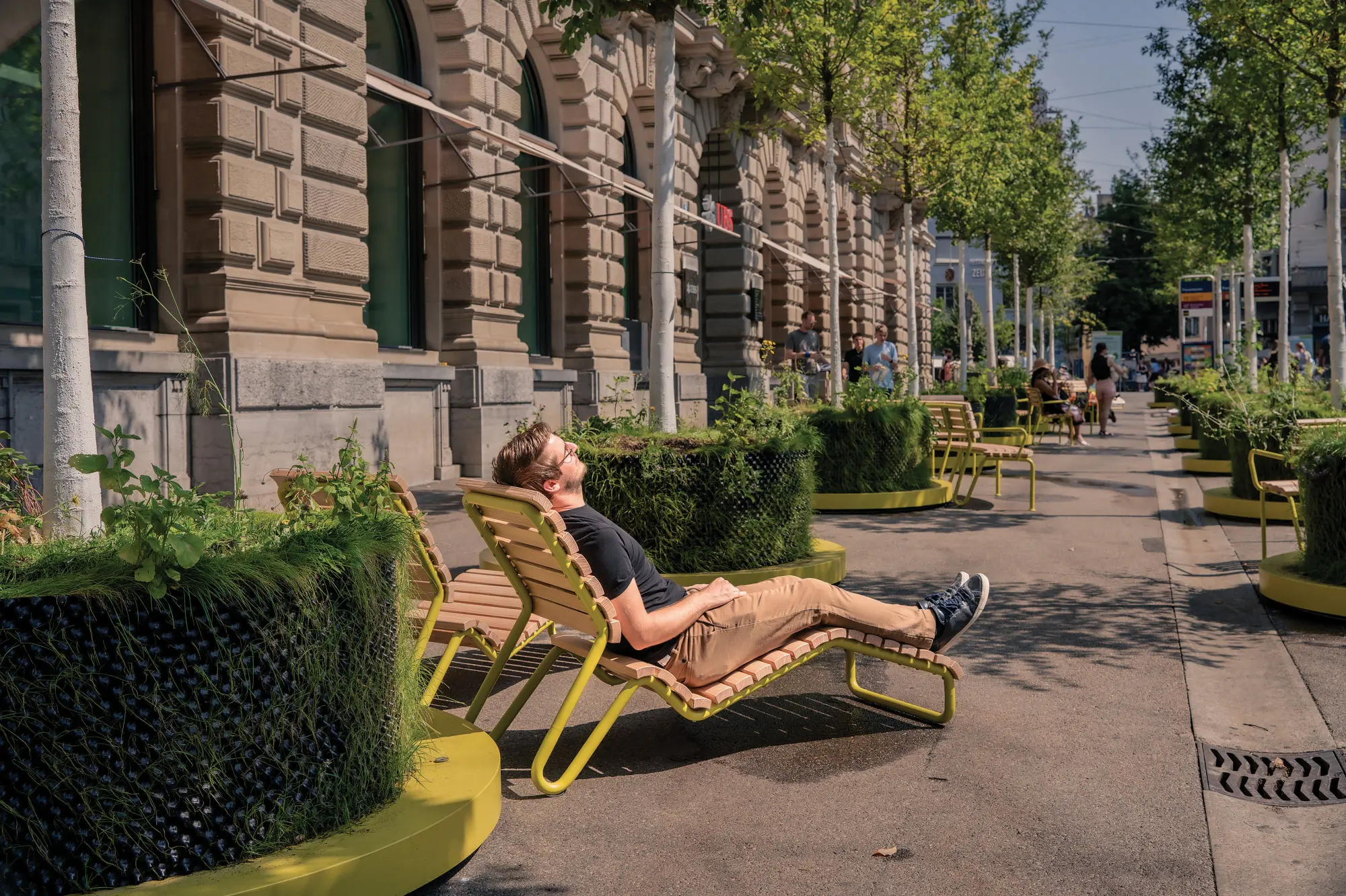 Liegestuhl aus mobilen Strassenmöbeln vor historischer Fassade in Zürich, Passant entspannt in der Sonne.