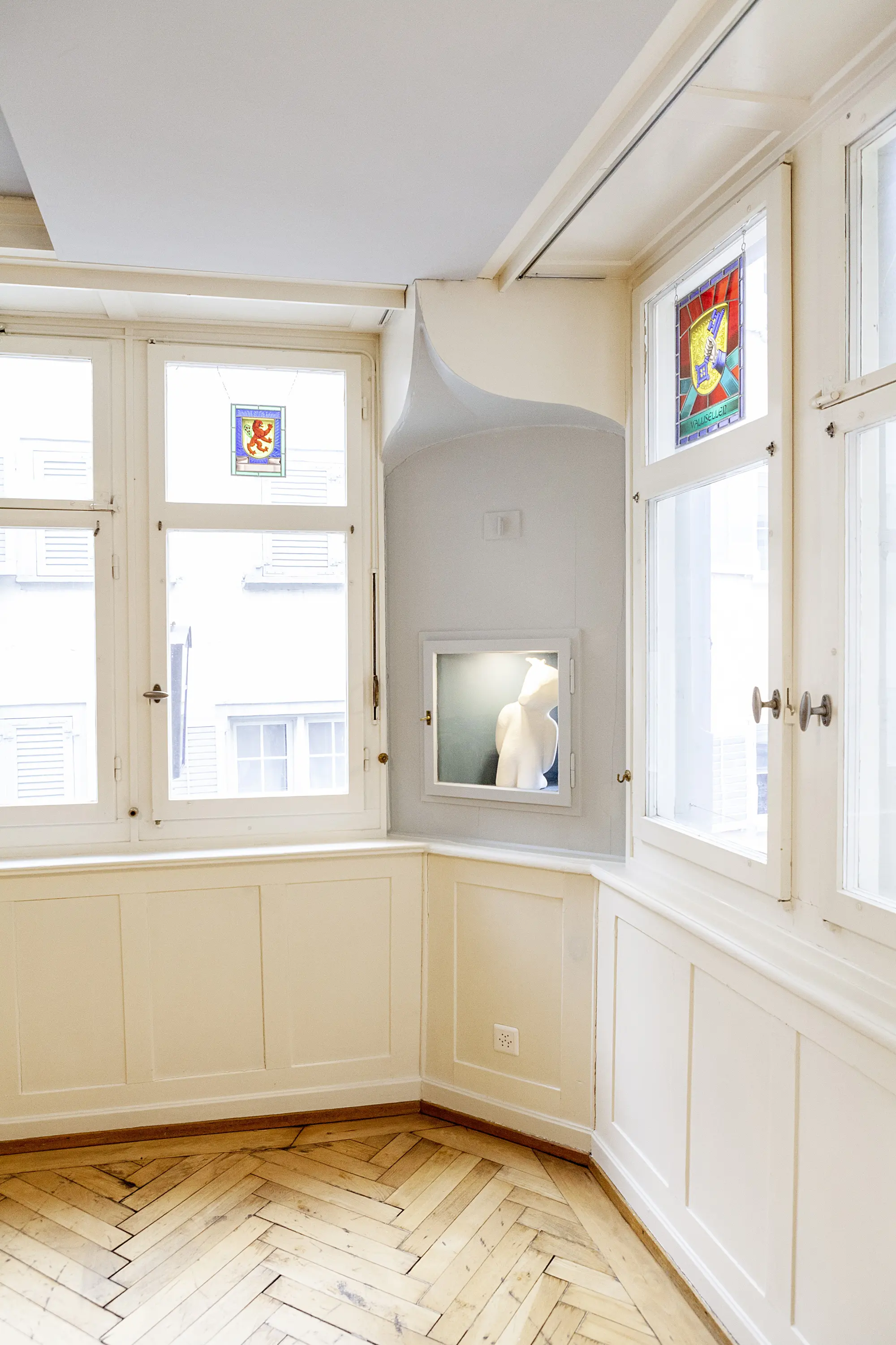 Helle Raumecke mit einem Kunstobjekt in der Wand, Bleiglasfenstern und Fischgrätparkett.