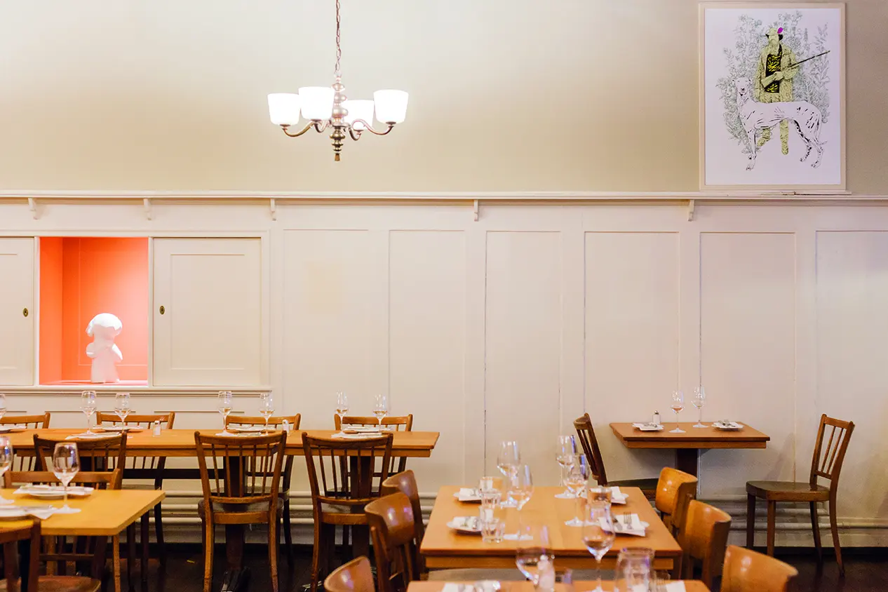 Heller Gastraum eines Restaurants mit gedeckten Holztischen, Holzstühlen, Kronleuchter und Kunstobjekten.