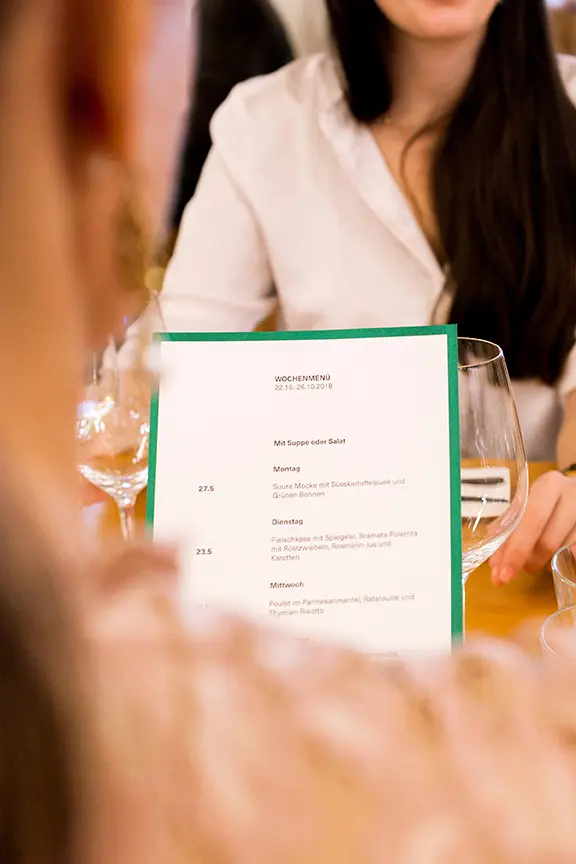 Gast hält eine grün gerahmte Wochenmenü-Speisekarte an einem gedeckten Restauranttisch.